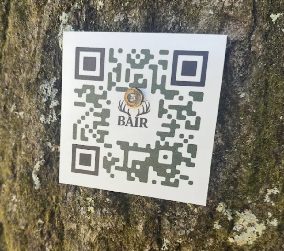 Close-up of a white QR sign with the Bair name and antler logo mounted on a tree.