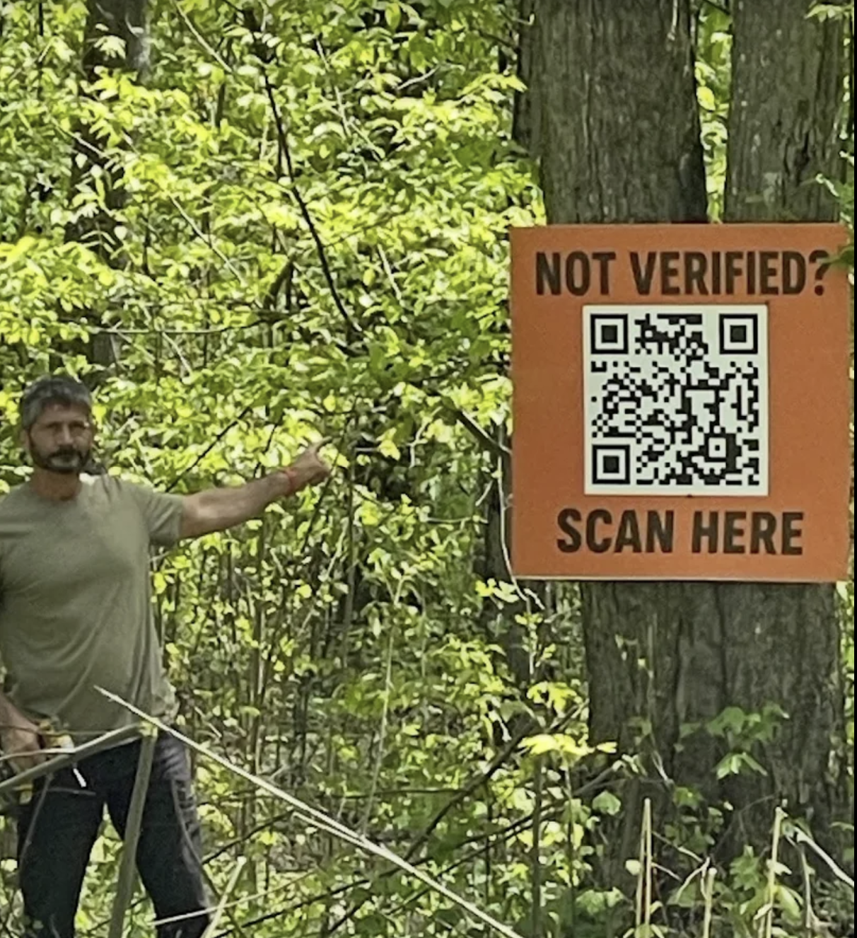 Orange QR sign mounted on a tree reading Not Verified? Scan Here, with a person standing beside it.